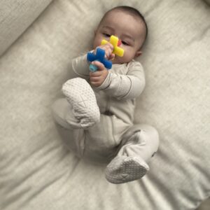 A baby in a gray onesie lying on a cushioned surface, holding a colorful toy with both hands and feet in the air.