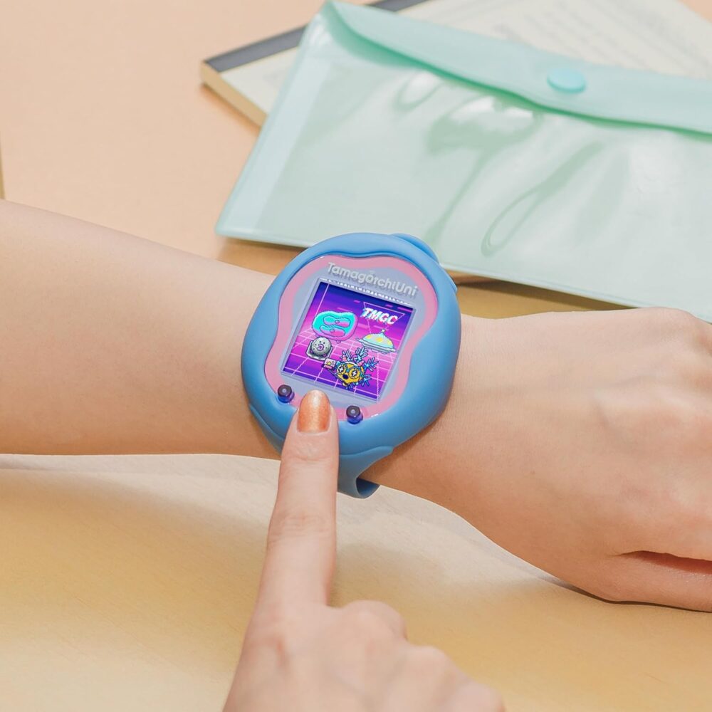 A person interacts with a blue Tamagotchi smartwatch displaying a colorful screen, while resting their hand on a light-colored surface. A translucent green envelope is partially visible in the background.