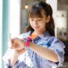 A young girl in a denim jacket looks at her wrist wearing a Tamagotchi Uni smartwatch in a brightly lit room.