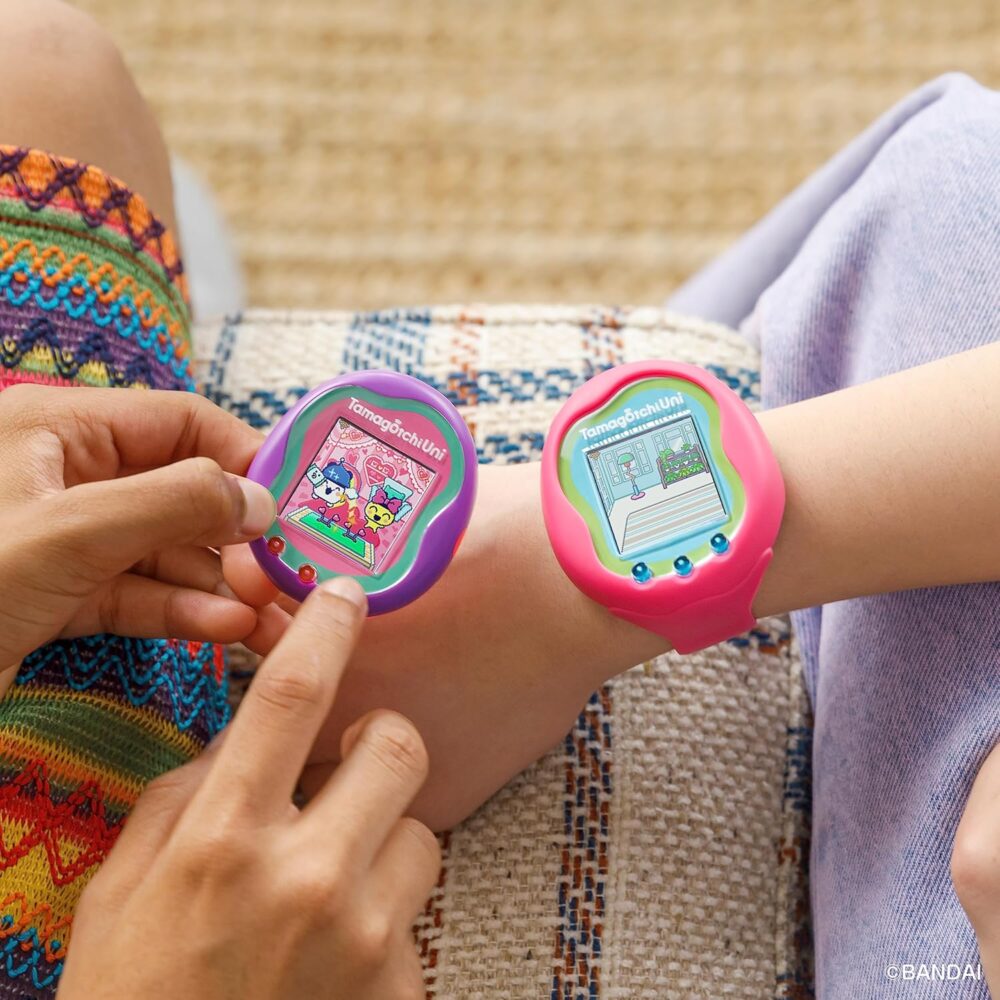 Two people interacting with pink Tamagotchi Uni wearable devices on their wrists, one person using finger to touch the screen. Both devices display digital pets.