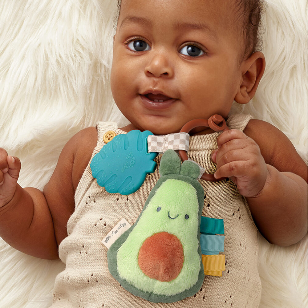 A baby holding an Itzy Ritzy - Itzy Pal - Plush Pal Infant Toy with Silicone Teether, lying on a white furry blanket, wearing a beige outfit.