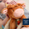 A baby holds a plush cow toy with a label reading "SNUGGLY SOFT LOVEY." The toy has brown fur, embroidered eyes, and a soft fabric underside with colorful ribbons.