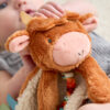 A close-up shows a baby holding a brown, plush toy cow. The toy has a round face, small horns, and floppy ears. The baby’s hand is visible, gripping the toy’s head.