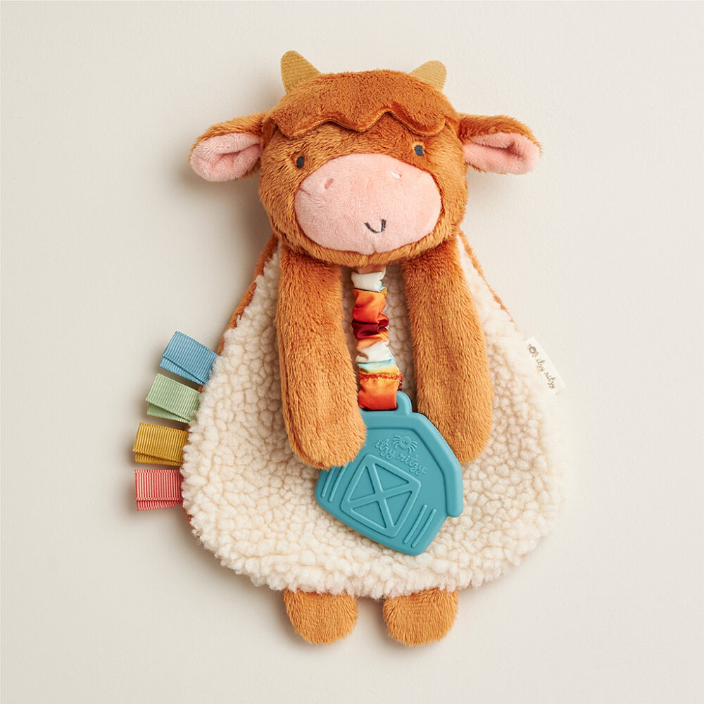 Plush toy of a smiling cow with brown fur, pink nose, and attached blue barn-shaped teether, adorned with colorful ribbon tabs on its left.