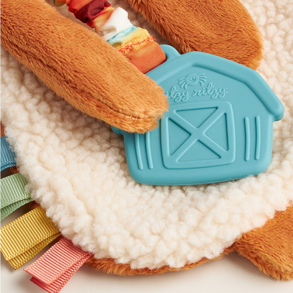 Close-up of a baby sensory toy with a blue silicone barn-shaped teether and colorful ribbons. The toy features soft, textured fabrics in various shades of brown and white.