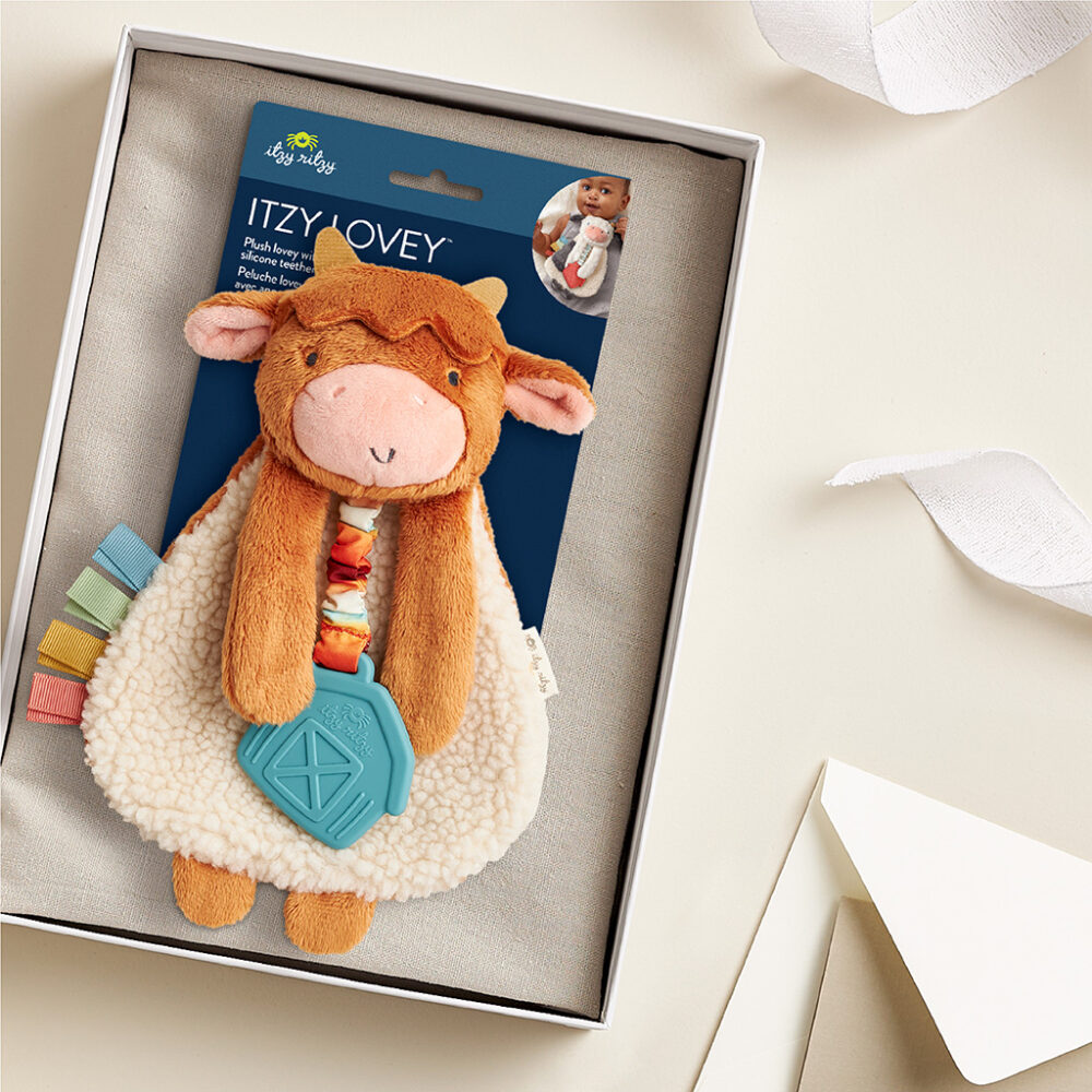 A plush toy packaged in a box labeled "Itzy Lovey" featuring a brown and white cow with a blue geometric teether and colorful ribbons attached. The background includes a white ribbon and an off-white envelope.