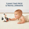 A baby lying on a beige carpet reaches toward a plush cow toy. Text above reads "Tummy Time Prop & Travel Linkmate.