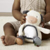 A baby in a beige knitted outfit plays with a plush cow toy while sitting on a cream-colored rug.