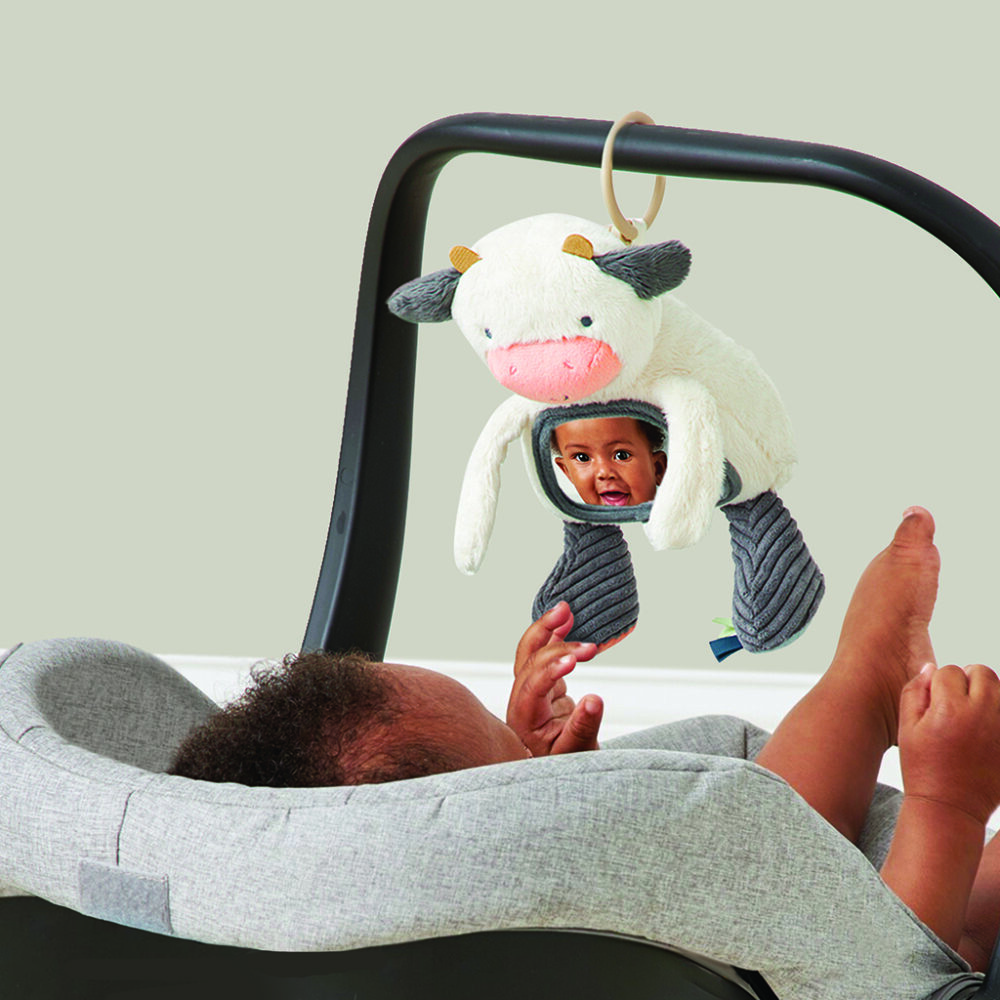 A baby lying in a gray car seat reaches up to touch a plush cow toy with a mirror and teething elements hanging above them.