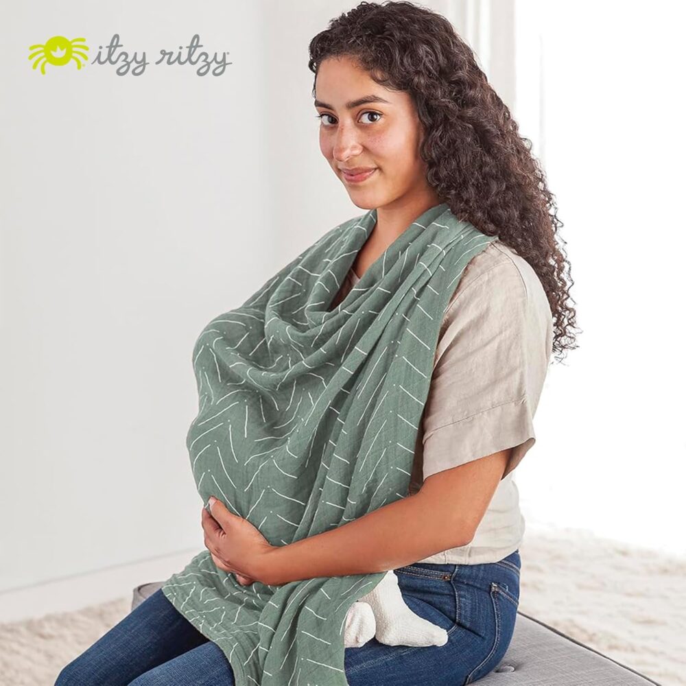 A woman sits on a cushioned surface, breastfeeding her baby under a light green striped Itzy Ritzy Breastfeeding Boss - a multitasking must-have for nursing, swaddling, and more. The Itzy Ritzy logo is clearly visible in the top left corner.