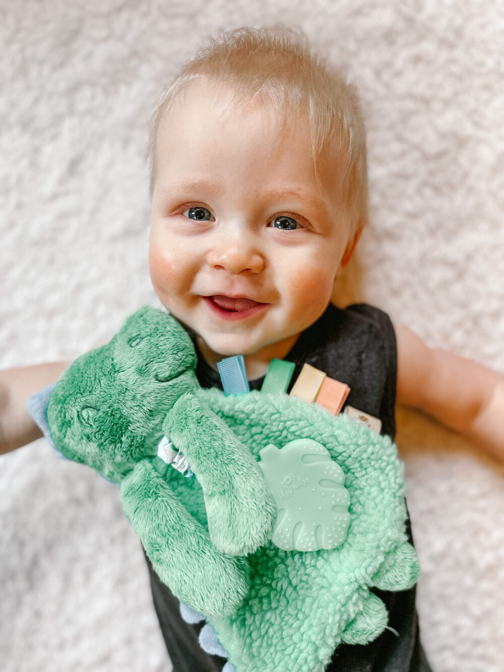 A baby lies on a soft surface, smiling and holding a green stuffed toy with colorful tags.