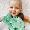 A baby lies on a soft surface, smiling and holding a green stuffed toy with colorful tags.