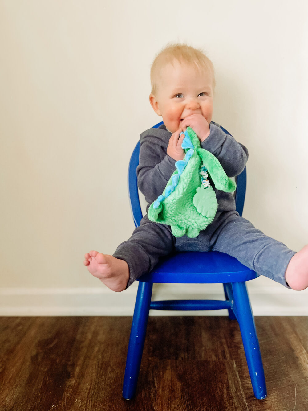 A baby sits on a blue chair, wearing a grey outfit and chewing on a green blanket, with feet playfully off the edge of the chair.