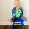 A baby sits on a blue chair, wearing a grey outfit and chewing on a green blanket, with feet playfully off the edge of the chair.