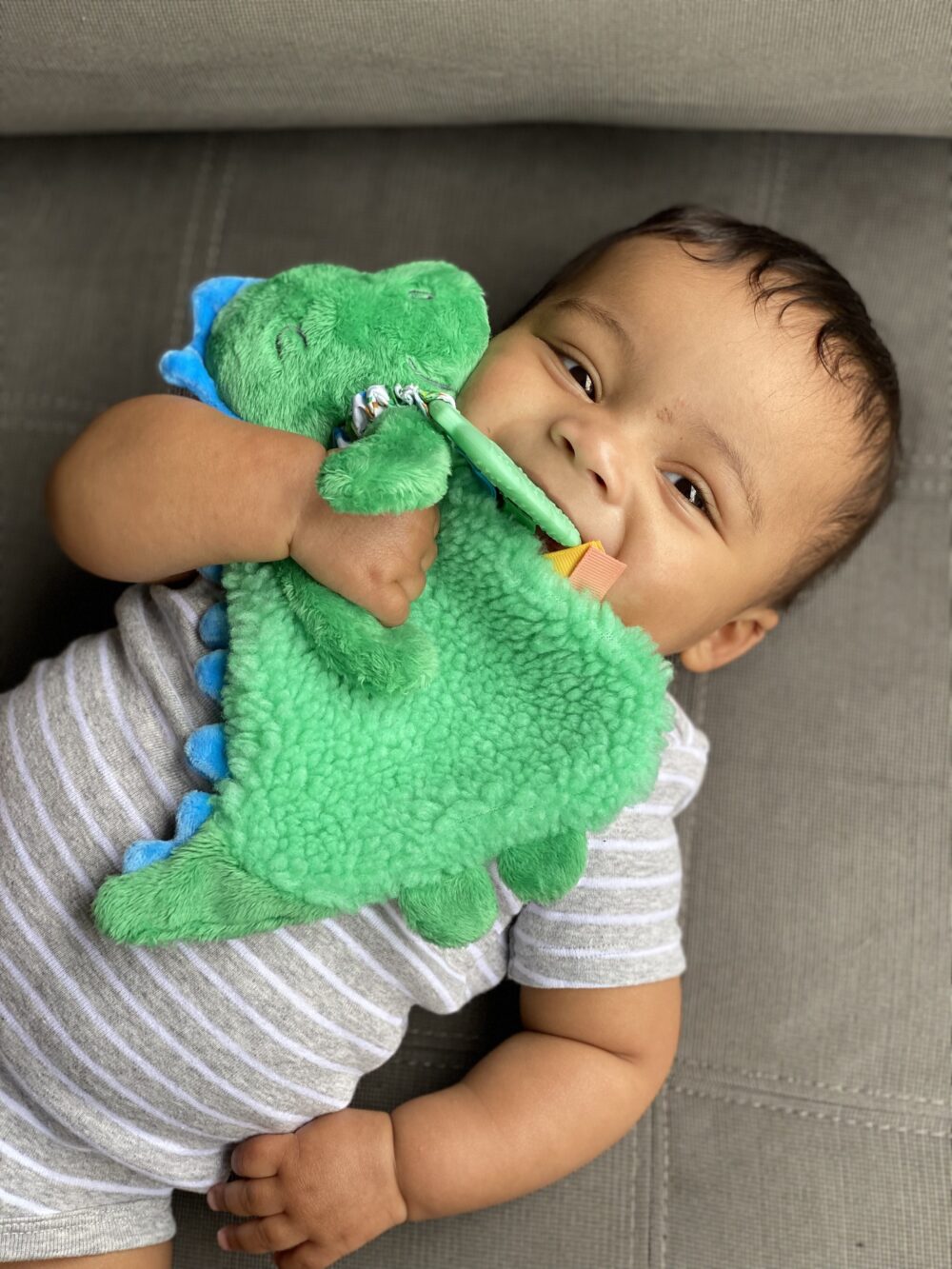 A baby lies on a grey cushion, holding and chewing on a green plush dinosaur toy while wearing a grey and white striped onesie.