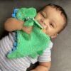 A baby lies on a grey cushion, holding and chewing on a green plush dinosaur toy while wearing a grey and white striped onesie.