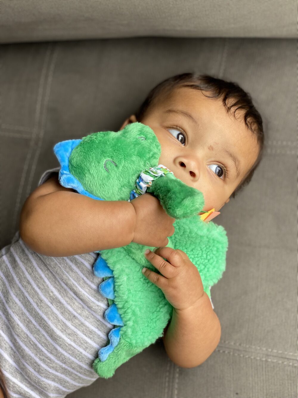 A baby lies on a gray surface, wearing a gray and white striped onesie, holding a green dinosaur plush toy close to their face.