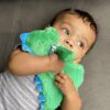 A baby lies on a gray surface, wearing a gray and white striped onesie, holding a green dinosaur plush toy close to their face.