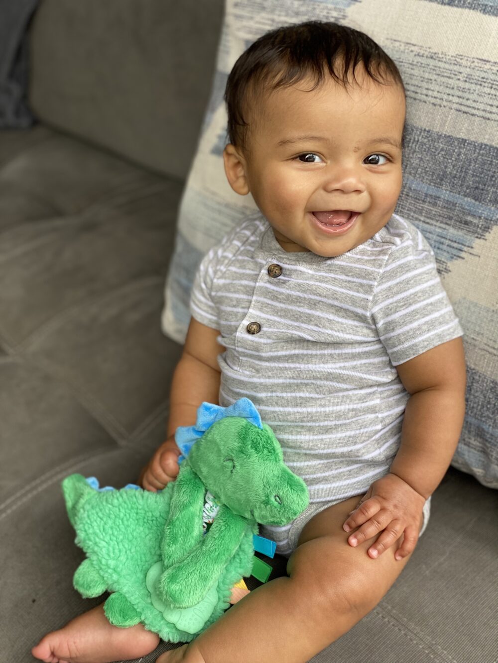 A baby with dark hair is sitting on a gray couch, wearing a striped onesie. The baby is smiling and holding a green plush dinosaur toy.