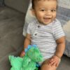 A baby with dark hair is sitting on a gray couch, wearing a striped onesie. The baby is smiling and holding a green plush dinosaur toy.