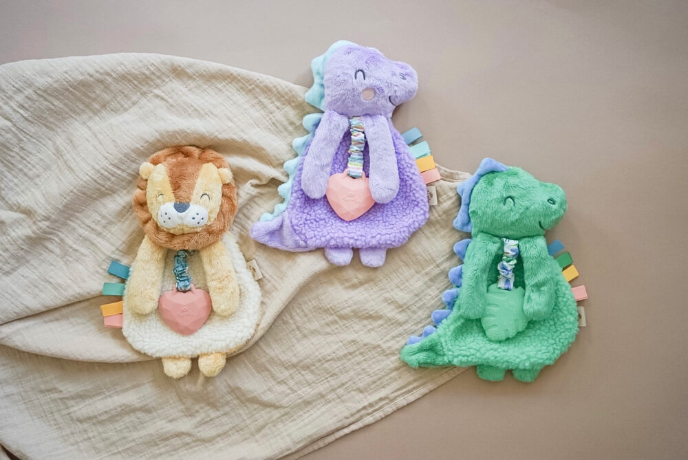 Three plush animal toys—a lion, a purple dinosaur, and a green dinosaur—lie on a soft beige blanket. Each toy holds a pink heart and has colorful tags on their backs.