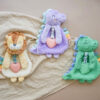 Three plush animal toys—a lion, a purple dinosaur, and a green dinosaur—lie on a soft beige blanket. Each toy holds a pink heart and has colorful tags on their backs.