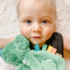 A baby with light hair lies on a white blanket, holding a green plush toy. The baby is wearing a dark outfit and looking directly at the camera.