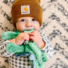 A baby wearing a brown beanie and a checkered shirt holds a green plush toy and looks at the camera, standing in front of a patterned background.