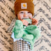 A baby, wearing a brown beanie and checkered outfit, smiles while holding and chewing on a green stuffed animal, standing in front of a patterned background.