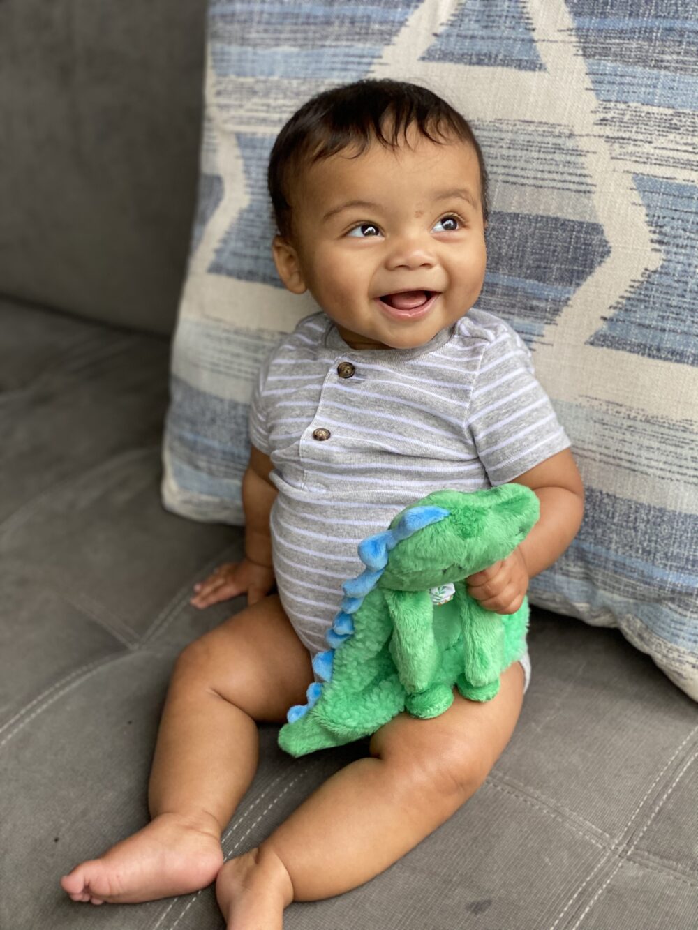 A smiling baby sits on a couch holding a green dinosaur plush toy, with a patterned pillow behind them.