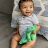 A smiling baby sits on a couch holding a green dinosaur plush toy, with a patterned pillow behind them.