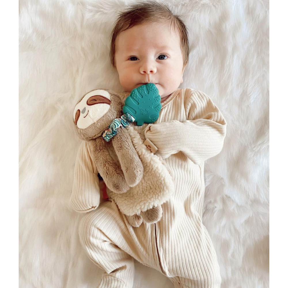 A baby dressed in a beige onesie lies on a white furry blanket, holding a plush sloth toy, and sucking on a green pacifier.