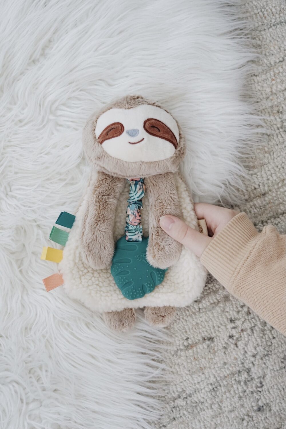 A hand holds a plush toy sloth with a white furry body and brown face, lying on a white fur rug. The toy features a teething ring and colorful fabric tags.