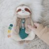 A hand holds a plush toy sloth with a white furry body and brown face, lying on a white fur rug. The toy features a teething ring and colorful fabric tags.