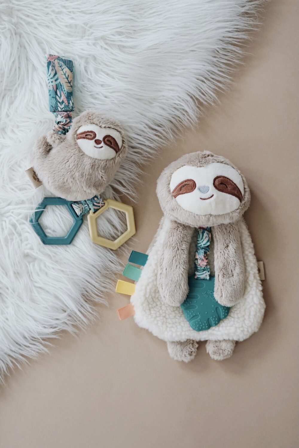 Plush sloth toys with hexagonal teething rings on a light surface; one smaller toy has two rings, and a larger toy has three attached fabric tags.