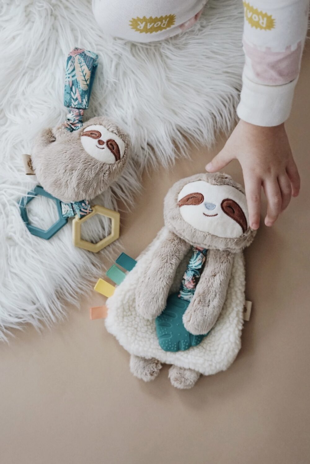 A child’s hand reaches for a plush sloth toy lying on a rug. The sloth toy has a smiling face and attached colorful ribbons. Another plush sloth toy with a plastic teether and geometric shapes lies nearby.