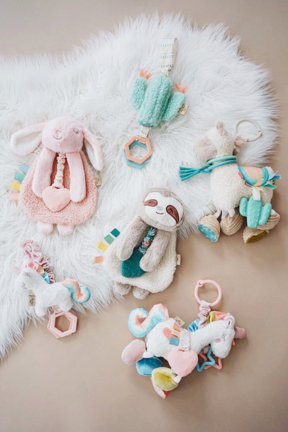 A variety of colorful plush baby toys including a pink rabbit, green sloth, llama, and unicorn are laid out on a white, furry surface.