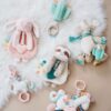 A variety of colorful plush baby toys including a pink rabbit, green sloth, llama, and unicorn are laid out on a white, furry surface.