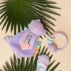 A plush purple dinosaur toy, a baby teething ring, and a baby mitten with rainbow designs are placed on green palm leaves set against a beige background.