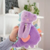 A hand is holding a purple stuffed toy shaped like a dinosaur with a small orange heart on its belly. The background shows white shelves with books and a plant.