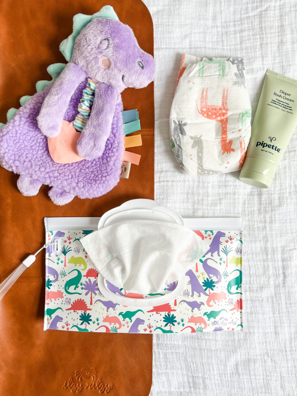 A flat lay of baby items: a purple dinosaur plush toy, a diaper with animal prints, a tube of pipette baby balm, a pack of wipes, and a brown changing pad all placed on a white cloth.