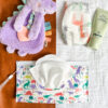 A flat lay of baby items: a purple dinosaur plush toy, a diaper with animal prints, a tube of pipette baby balm, a pack of wipes, and a brown changing pad all placed on a white cloth.