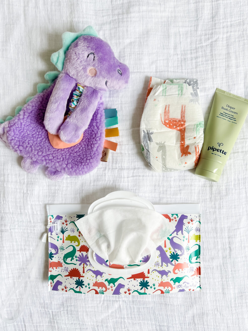 A purple plush dinosaur toy, a diaper with dinosaur prints, a tube of Pipette baby cream, and a pack of dinosaur-themed baby wipes on a white fabric background.