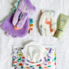 A purple plush dinosaur toy, a diaper with dinosaur prints, a tube of Pipette baby cream, and a pack of dinosaur-themed baby wipes on a white fabric background.