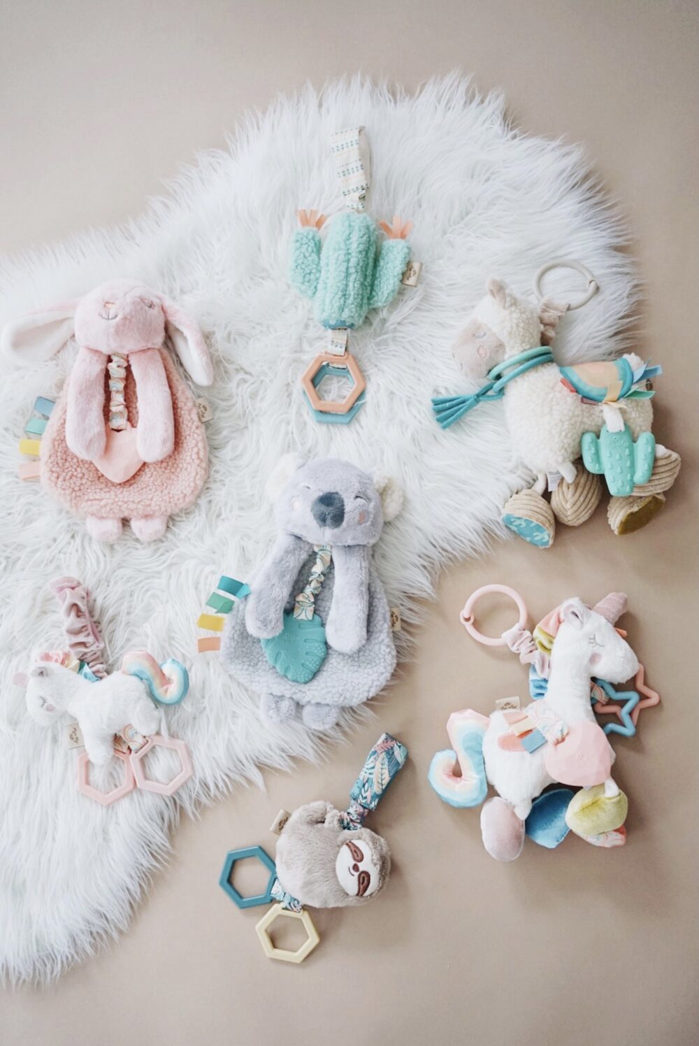 Plush baby toys and teethers, including a bunny, lamb, unicorn, bear, and sloth, are arranged on a soft white rug on a beige background.