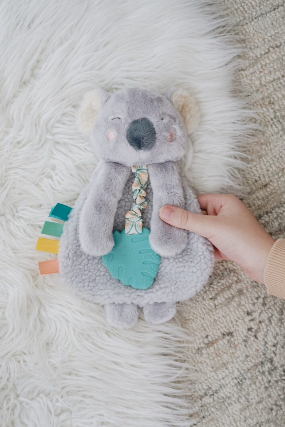 A hand holding a plush koala toy with a teal teether attached to its front and colorful fabric tabs on its side, lying on a fluffy white surface.