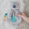 A hand holding a plush koala toy with a teal teether attached to its front and colorful fabric tabs on its side, lying on a fluffy white surface.