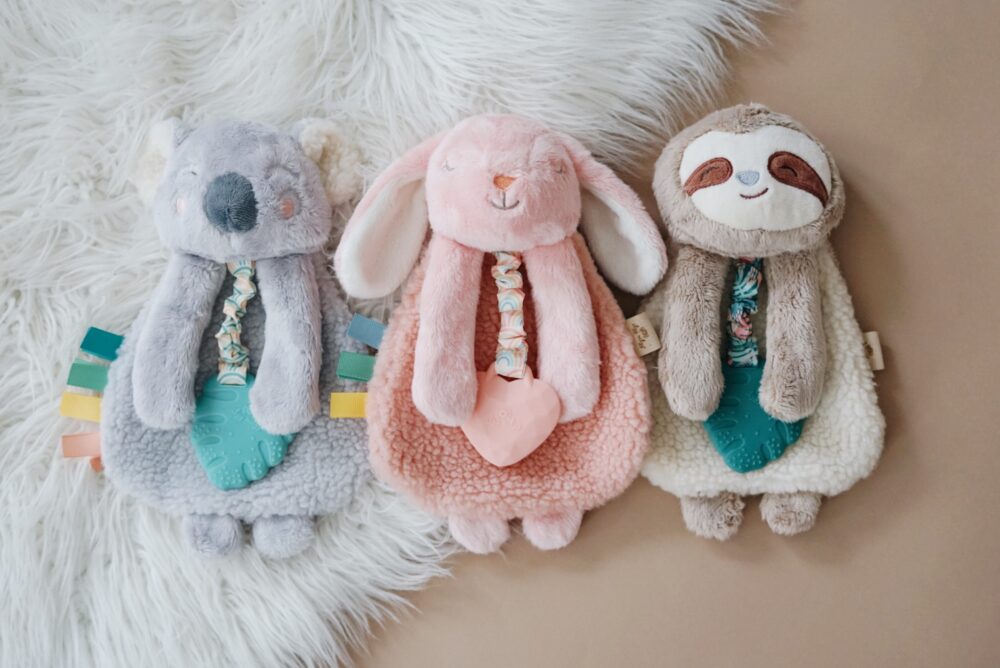 Three stuffed animals lying side by side on a soft surface: a grey koala, a pink bunny, and a tan sloth, each with a bib and teething toy attached.