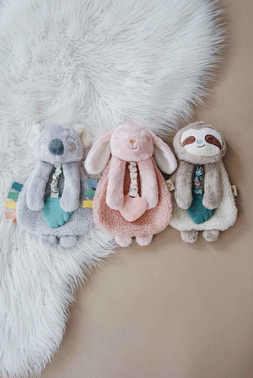 Three plush toys, including a koala, a bunny, and a sloth, lie on a white faux fur rug and beige background. Each toy holds a teething ring and has adorably embroidered details.