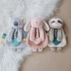 Three plush toys, including a koala, a bunny, and a sloth, lie on a white faux fur rug and beige background. Each toy holds a teething ring and has adorably embroidered details.